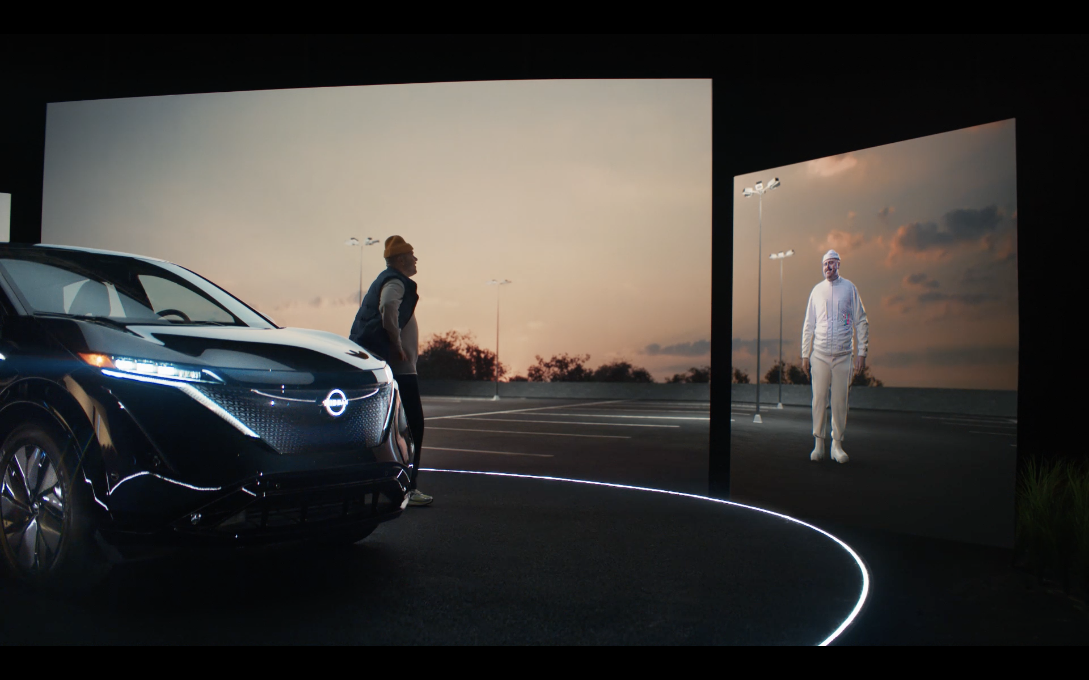 A person standing near a Nissan ARIYA electric vehicle, interacting with a life-sized avatar of themselves projected on a screen.